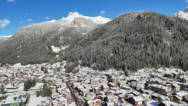 Canazei, Italy: Aerial view of ski resort in snowy Dolomites. Charming town landscape with chalets and hotels surrounded by snow-capped peaks, sunny day, clear blue sky
