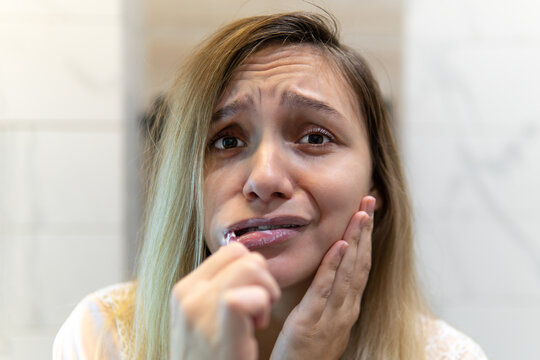 sad girl with a toothbrush in her mouth looks in the mirror. The concept of toothache and suffering. A young woman is brushing her teeth.