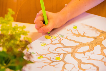 Close-Up of Hand Writing on Illustrated Family Tree Diagram with Green Marker, Surrounded by Decorative Leaves, Showcasing Ancestral Connection and Genealogy Research in Bright Indoor Setting