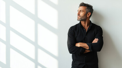 Middle-aged man in black shirt poses thoughtfully by a sunlit wall, casting geometric shadows. He exudes a calm and introspective mood.