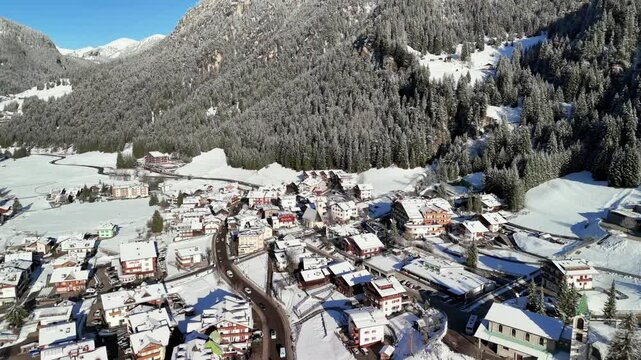 Canazei, Italy: Aerial view of ski resort in snowy Dolomites. Charming town landscape with chalets and hotels surrounded by snow-capped peaks, sunny day, clear blue sky
