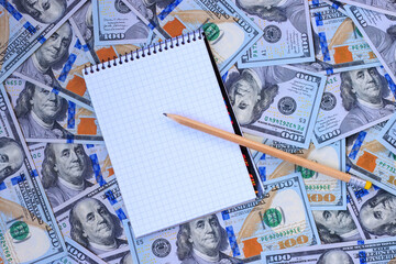 One hundred dollar bills in the background, a notebook, and a pencil, personal finance concept.