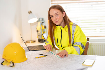 Young woman site manager in safety gear planning construction project indoors with tools and laptop in bright workspace