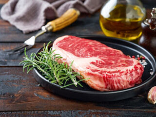 Raw beef steak on black plate with pepper, salt, rosemary and olive oil on black stone table with ample copy space. Dark background. Copy space. Soft focus.