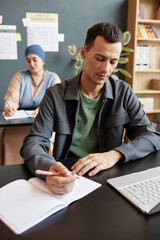Vertical shot of adult multiracial male student focused on writing task sitting at desk in study...