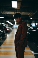 A young man in a brown suit standing confidently in a dimly lit underground parking lot