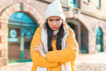 Young woman in yellow coat frowning with arms crossed in a winter urban setting