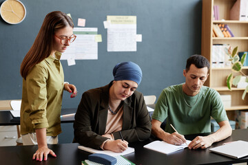 Medium shot of adult man and Muslum woman writing in notebooks sitting at desk with teacher...