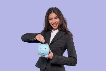 Young woman in business attire happily saving coins in a blue piggy bank against a pastel background