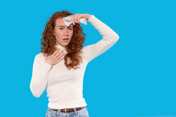 Young woman with curly hair wipes forehead while feeling unwell against bright blue background