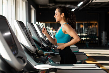 Fototapeta premium Fit European woman in sportswear running jogging at treadmill and smiling, enjoying morning workout in gym, side view