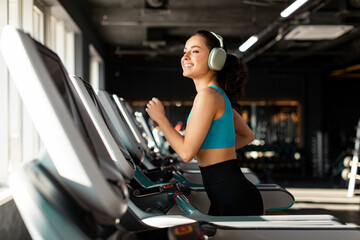 Happy European woman in sportswear and wireless headphones doing hard cardio workout on treadmill,...