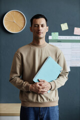 Vertical portrait shot of happy mixed raced male student posing with exercise book in hands while standing against blue wall in school classroom, copy space