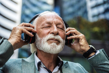 Senior businessman, headphones and listening to music in city for travel, motivation and happy mindset. Mature men on an urban street with buildings background while streaming podcast or audio outdoor