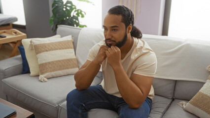 Obraz premium Young man with beard sitting thoughtfully in modern living room interior with cushions and plants, embodying a calm and relaxed atmosphere at home.