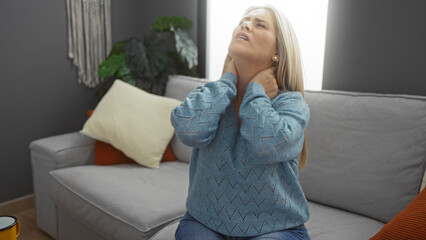 Woman experiencing discomfort in neck sits on gray sofa in cozy living room with stylish decor and soft lighting, conveying a sense of pain and stress at home.