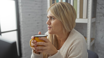 Woman holding mug indoors in office setting with blonde hair, wearing sweater, looking thoughtful, sitting near window with books on shelves in the background.