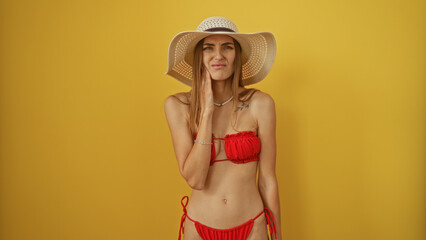 Woman experiencing toothache in a red bikini and wide-brimmed hat, isolated against a yellow...