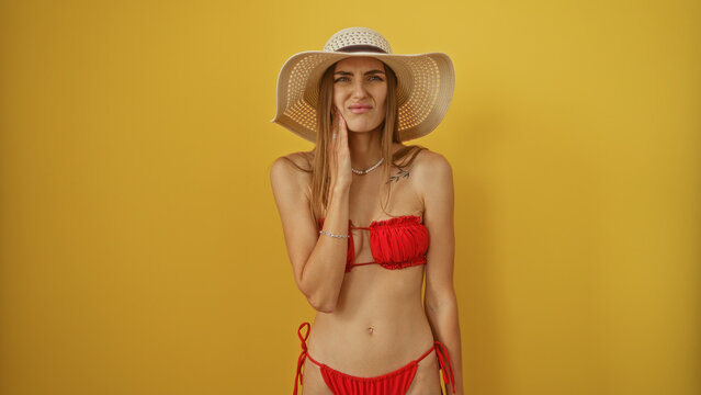 Woman experiencing toothache in a red bikini and wide-brimmed hat, isolated against a yellow background, expressing discomfort while touching her cheek.