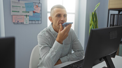 Young man using smartphone for voice command in modern office setting, surrounded by computer, plants, and strategic planning notes