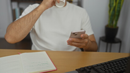 Fototapeta premium Hispanic man in an office sipping coffee while using a smartphone, with hands visible near a notebook on the desk.