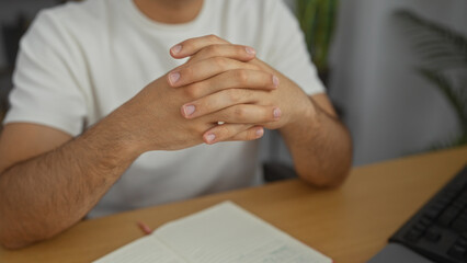 Obraz premium Male adult person in an indoor office, hispanic man with folded hands sitting at a desk with an open notebook and a computer keyboard.