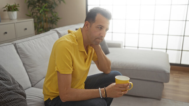 Hispanic man sitting in a living room holding a yellow mug with a thoughtful expression