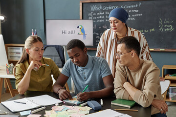 Medium shot of young Black man writing word on post it note for playing vocabulary game with teacher assisting student, while studying in collaborative activity for adult learners