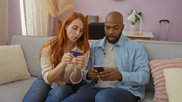Interracial couple sitting on a couch at home discussing finances with a credit card and smartphone, displaying a relaxed family-oriented setting with a modern interior