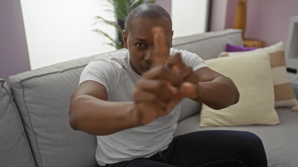 Fototapeta premium Young man sitting on sofa in contemporary living room showcasing a relaxed atmosphere, while playfully gesturing at the camera with a neutral expression in his stylish apartment.
