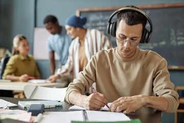 Medium shot of multiracial male student in headphones concentrated on completing task developing listening skills while sitting at table in language school for adult learners, copy space