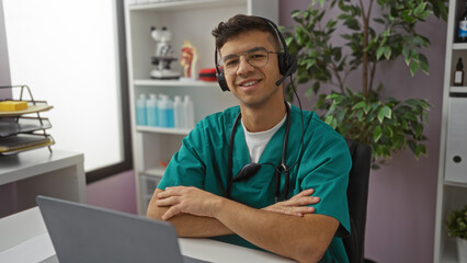 Young hispanic man in medical scrubs with headset and glasses, arms crossed, smiling in a hospital...