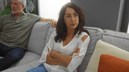 Obraz premium Woman with crossed arms sits on couch next to man in living room depicting couple in disagreement during tense moment indoors
