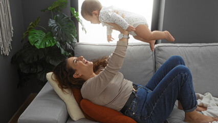 Naklejka premium Woman smiling while lifting baby in cozy living room, expressing joyful family love and connection, interior scene shows warm home environment, emphasizing mother-daughter bond.