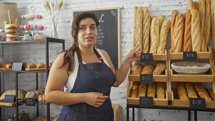 Young woman with brunette hair in a bakery shop pointing at various bread loaves and prices on display shelves wearing a blue apron.