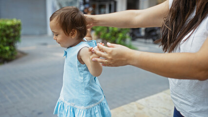 Obraz premium Woman guiding toddler girl outdoors on urban street holding her hands wearing blue dress with city background