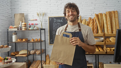 Handsome young man working in a bakery holding a paper bag, surrounded by various breads in an indoor shop setting.