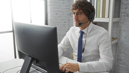 Young man working in office wearing headset and glasses focused on computer in modern indoor setting with bookshelf and window background showing professional environment