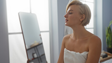 Woman relaxing in a spa salon with a towel wrapped around her body, looking into a mirror in a serene wellness center with natural light and indoor plants