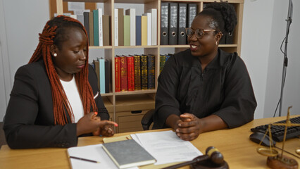 Two african american women, a lawyer and a judge, discussing documents in an office with legal books in the background.