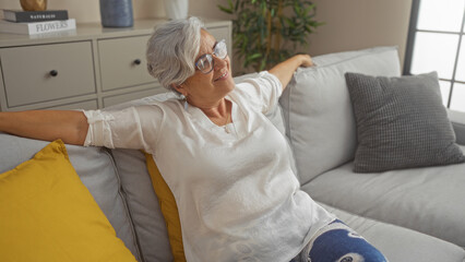 Fototapeta premium Mature woman relaxing on a sofa in a cozy living room, enjoying a serene moment in her home environment, surrounded by cushions and decor.