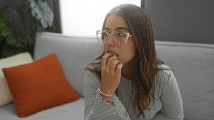 Fototapeta premium Woman indoors with glasses sitting thoughtfully on a couch in a cozy living room, showcasing a relaxed and modern home setting.