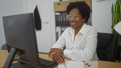 African american woman with curly hair and glasses working on a computer in an office, smiling...