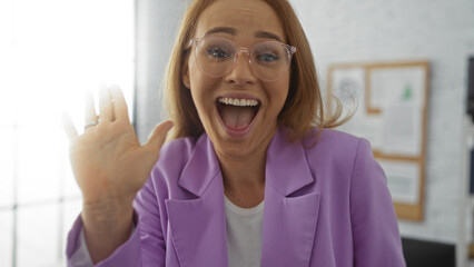 Woman with glasses waving joyfully in a bright office setting, wearing a purple blazer, showcasing...