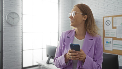 Woman smiling in modern office holding phone, wearing glasses and purple blazer, stands near large window and clock in bright, organized workplace.