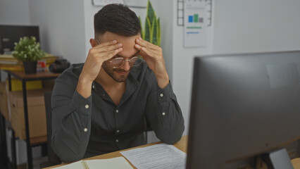 Naklejka premium Handsome young hispanic man with beard working in an indoor office, visibly stressed while looking at documents on his desk.