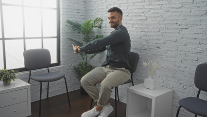 Handsome young hispanic man with a beard sitting in a modern indoor waiting room, stretching his arms with a smile.