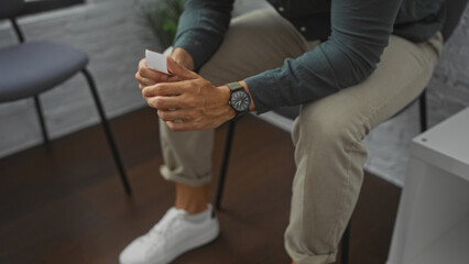 Hispanic man sitting in an indoor waiting room, holding a document, with hands visible and wearing a watch, suggesting a patient or applicant in a lobby setting.