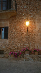 Warm streetlamp illuminates potted pink flowers against a rustic stone wall with a black iron balcony in the quaint village setting.