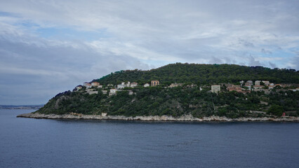 Fototapeta premium Scenic view of coastal hillside with lush greenery and houses overlooking calm sea under cloudy sky.
